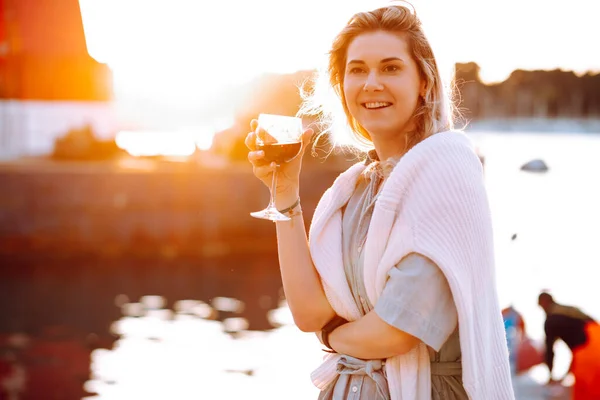 Portrait of smiling woman holding in hand wine glass and drinking red wine at sunset on blurred evening pier background. Walking along sea embankment with boats and yachts in resort city