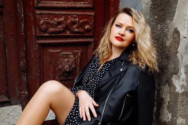 Portrait of beautiful blonde hair woman with bright makeup and perfect curls sitting and posing on old building background in casual style clothes. Fashion outfit for street walking on cool day — Photo