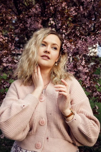Portrait of beautiful blonde hair woman with gentle makeup and perfect curls posing on blooming lilac background in pink knitted cardigan. Fashion outfit for street walking on spring city park — Zdjęcie stockowe