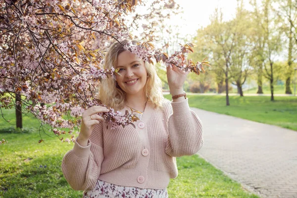 Portrait of smiling beautiful blonde hair woman in pink knitted cardigan holding branches of blooming lilac on sunny day, nature background. Fashion outfit for street walking on spring city park — Foto Stock