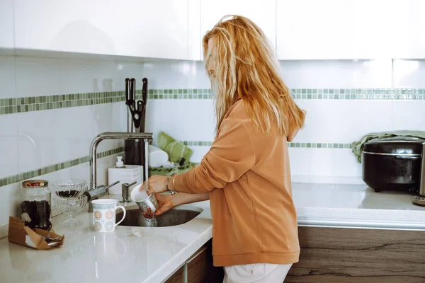 Blonde young woman in home clothes wash mugs in sink under running water after eating and tidy up modern kitchen side view. Housewife clean up house and wash all dirty dishes. Clean house concept