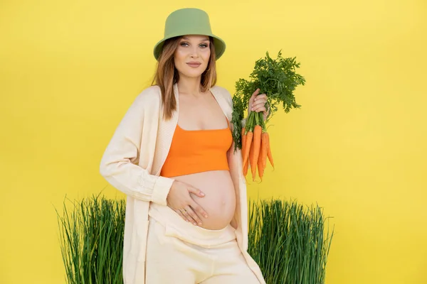 Woman with bare belly in late pregnancy on grass decoration holding fresh carrot bunch in casual clothes, yellow background. Portrait of young pregnant lady expecting baby. Healthy pregnancy concept — Stock Fotó