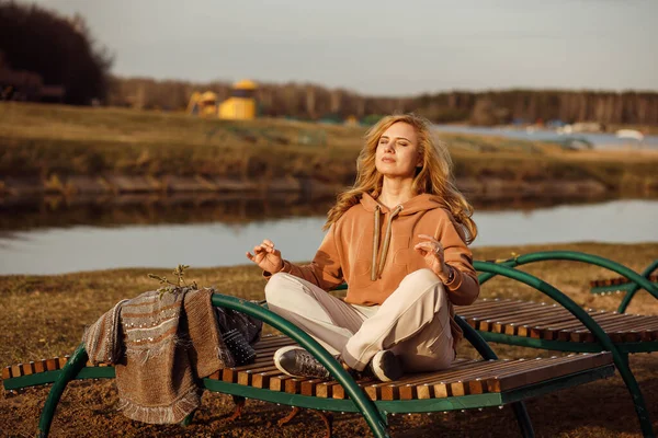 Красива блондинка сидить на лавці поруч з берегом річки в холодний сонячний день, розслабляючись, роздумуючи і насолоджуючись миром і свободою. Активність на відкритих вихідних, сільський фон природи — стокове фото