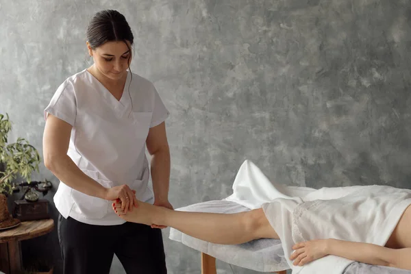 Остеопат в медицинском кабинете массирует ногу женщины, лежащей на больничном диване под белым покрывалом. Назначение и осмотр профессионального специалиста. Концепция лечения и лечения заболеваний — стоковое фото