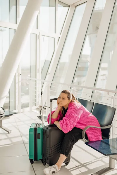 Tired passenger sitting on chair in light airport with panoramic windows, lying down on suitcases with clothing and waiting airplane arriving, flight delay. Young woman travel and relax on vacation — Stock Photo, Image