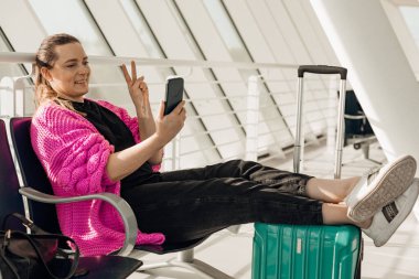 Portrait of smiling woman sitting, stretching legs, having video-call, holding smart phone in hand, taking selfie.