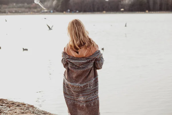 Вид сзади женщины с длинными волнистыми светлыми волосами в коричневой толстовке, длинном кардигане, стоящей на песчаном пляже реки. — стоковое фото