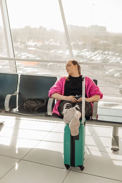 Portrait of tired woman wearing pink cardigan, sitting on chair, stretching legs on turquoise suitcase, sleeping.