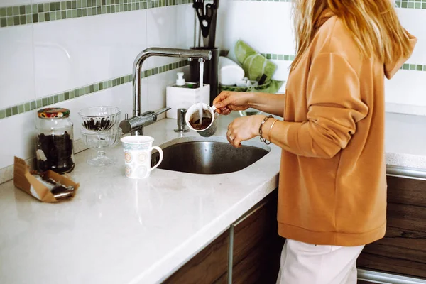Foto recortada de una mujer joven con el cabello ondulado y rubio con sudadera marrón, lavando la taza de café bajo agua abierta. — Foto de Stock