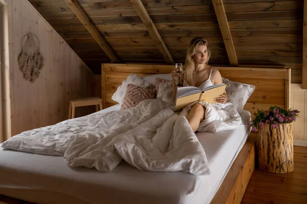 Portrait de jeune belle femme aux cheveux longs et luxuriants se relaxant au lit le matin avec un verre de vin et un livre. Repos à l'hôtel en montagne. Intérieur en bois du chalet. Week-end vacances. — Photo