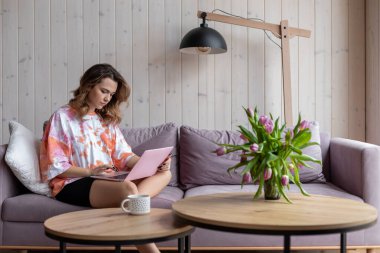 İnternette evde çalışıyorum. Rahat ev kıyafetleri içinde uzun saçlı, çekici, kahverengi saçlı bir kadın internette rahat bir koltukta oturuyor. Modern teknolojiler.