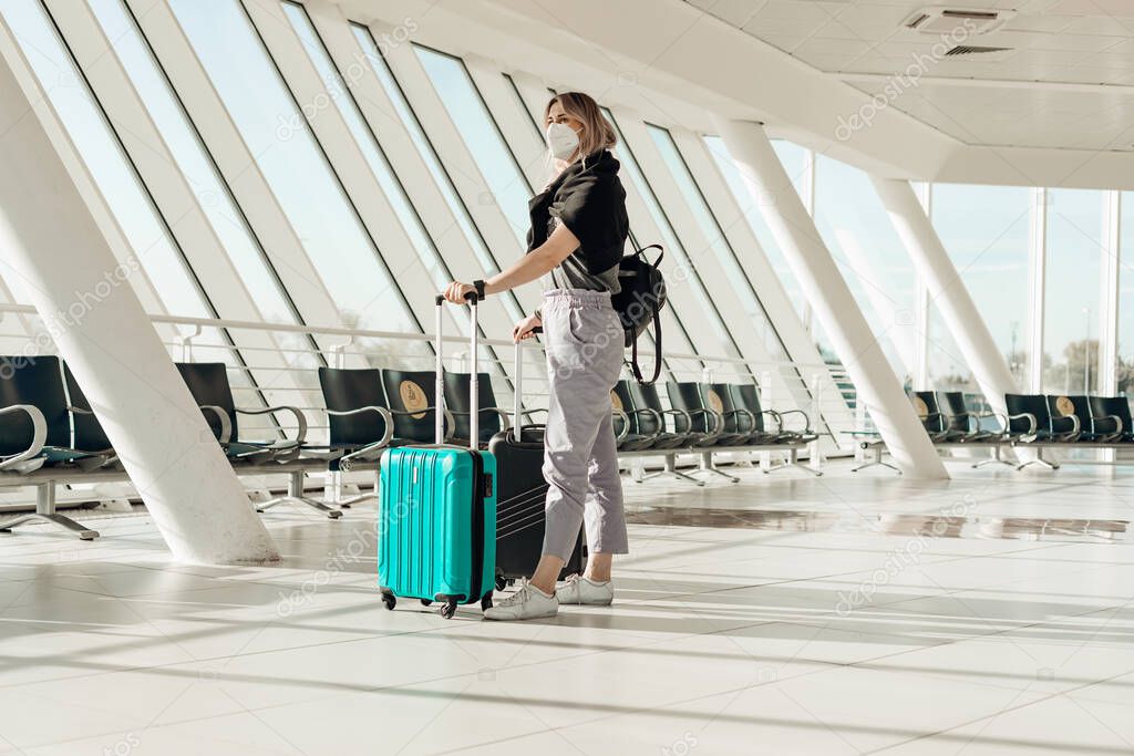 Portrait of woman wearing black T-shirt, grey trousers, disposable mask, standing, holding turquoise, black suitcases.