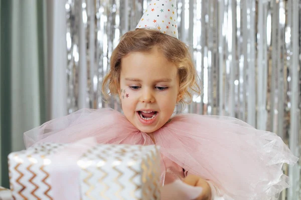 stock image Celebrating birthday. Happy festively dressed 4-5 - year - old girl with stars on her cheeks in party hat is holding gift box in her hands. Gifts for children.