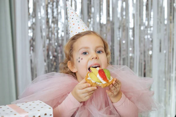 Happy 4-5-year-old girl in party hat in pink carnival dress is going to try cake with whipped cream and fruit, gift box is standing next to her on table. Birthday. Family holiday.