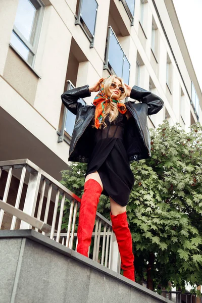 Uzun dalgalı sarı saçlı, kısa siyah elbiseli, deri ceketli, bina kenarında duran genç bir kadın..