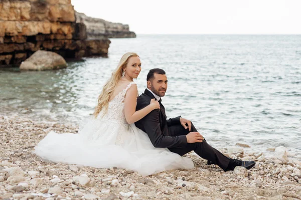 Retrato de noiva loira jovem em vestido branco e noivo morena em terno abraçando e sentado na costa do mar rochoso na Itália vista lateral, fundo da paisagem marinha. Casamento bonito e romântico, casal feliz — Fotografia de Stock