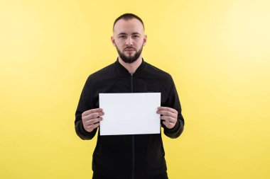 Ciddi sakallı iş adamı elinde beyaz boş bir kağıt parçasıyla ayakta duruyor, sarı arka plandaki kameraya bakıyor.