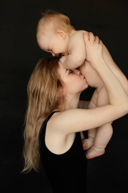 Siyah arka planda büyüyen ve tatlı, çıplak bir bebeği öpen genç bir annenin portresi. Mutlu ebeveyn anneliği seviyor. Anne ve küçük çocuk ilişkileri. Aile ve bebek bakımı kavramı.