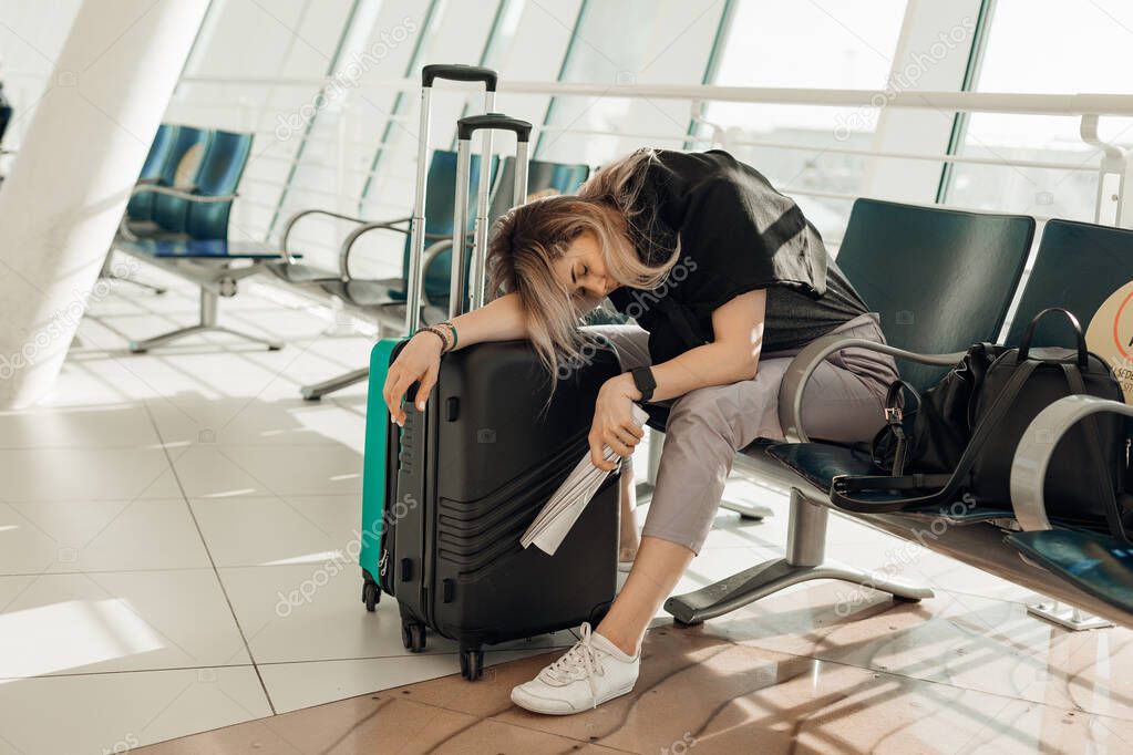 Tired passenger sitting on chair in modern light airport, lying down on suitcases with clothing and waiting airplane arriving, flight delay. Young woman fly on plane to travel and relax on vacation.