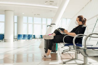 Siyah elbiseli bir kadın, tek kullanımlık beyaz bir maske, havaalanının ulaşım salonunda bavulların yanında oturan, dergi okuyan, uçuş uçaklarını bekleyen bir kadın. Kişisel sağlık güvenliği.