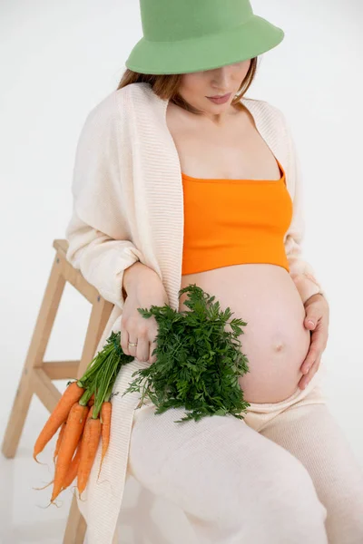怀孕后期赤身裸体的女人坐在椅子上 手里拿着胡萝卜束 头戴睡帽 背景是白色的 年轻孕妇期待小宝宝特写 — 图库照片