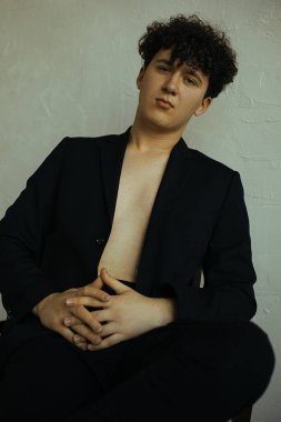 Kıvırcık siyah saçlı genç adamın dikey portresi çapraz el ele, kameraya, oturmaya, stüdyo çekimine bak. Retro tarzı. Çıplak vücudunda siyah ceketi olan çekici ve yakışıklı bir adam. Klasik duvar arkaplanı