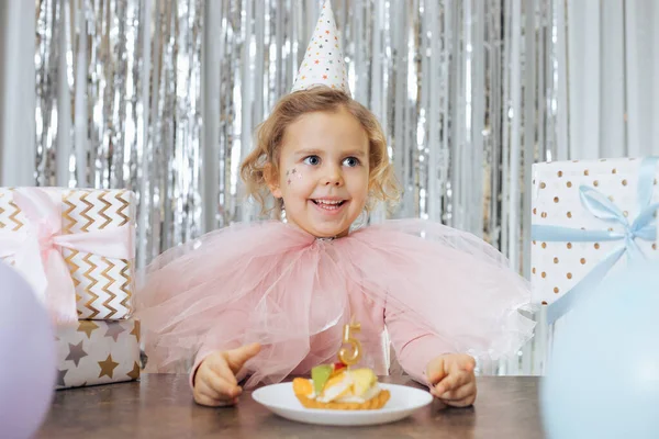 Happy moments. Childrens positive emotions. Smiling girl in festive outfit is sitting in front of birthday cake decorated with number five, surrounded gift boxes and balloons.