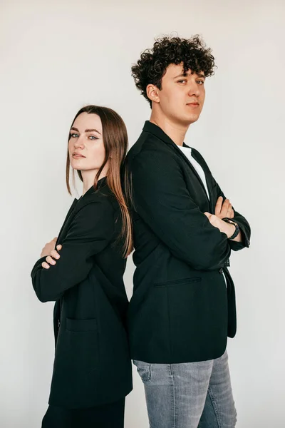 Vertical portrait of young attractive dark-haired man and woman in black clothes standing back to back together with crossed arms. Looking at camera. Studio shot on white isolated background.
