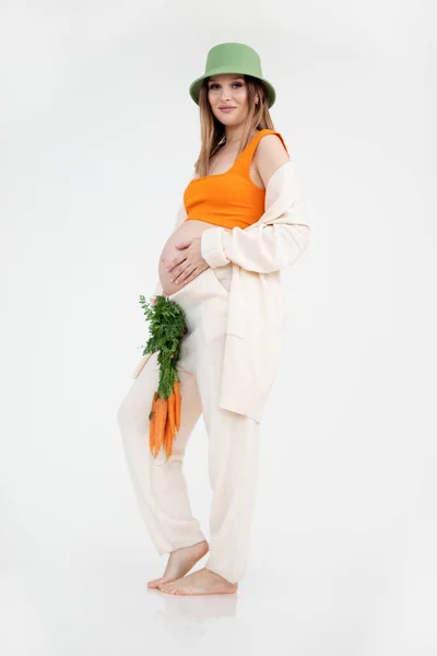 穿着时尚服装 头戴帽子 头戴白色背景胡萝卜 赤脚站立 健康饮食理念的孕妇 健康的新鲜蔬菜维生素营养食物 — 图库照片
