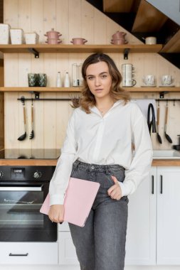 İç mimar. Beyaz gömlekli, uzun saçlı ve kot pantolonlu çekici bir kadın elinde dizüstü bilgisayarla mutfağın arka planına karşı duruyor. İnternette evde çalışıyorum. Yaşam biçimi. 