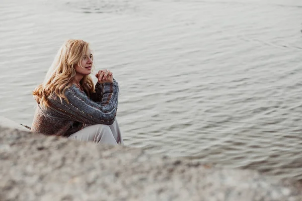 Portrait of young good-looking woman with long wavy fair hair wearing brown woolen cardigan, beige trousers, sitting at bank of river near concrete embankment, joining hands, interlacing fingers.