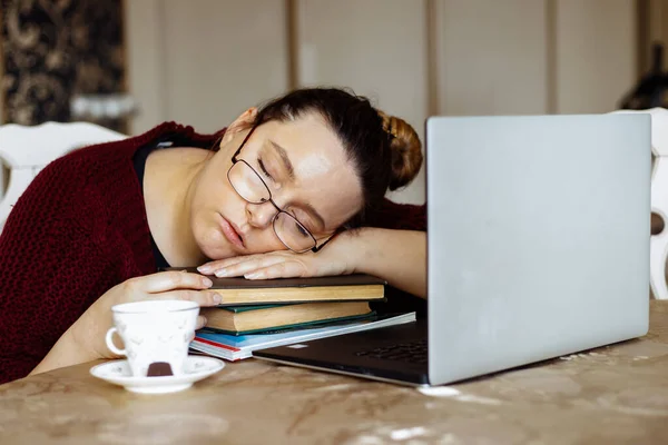 Portrait of exhausted middle-aged woman with long dark hair in bun wearing vinous sweater, glasses, sitting at table near cup of coffee, laptop, sleeping on crossed arms on pile of books. Hard work.