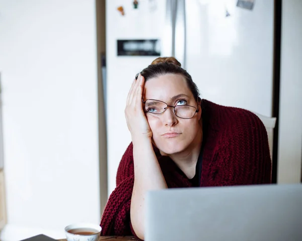 Portrait of middle-aged dissatisfied woman with dark hair in bun wearing vinous cardigan, glasses sliding up, leaning on table with elbow at home, looking up near laptop, cup of coffee, thinking.