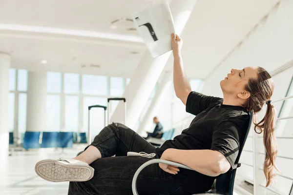 Side view of woman wearing black clothes, sitting on chair in transit lounge of airport, waving with magazine, suffering from heat, waiting for flight aircraft. Savings on air conditioning in Europe.