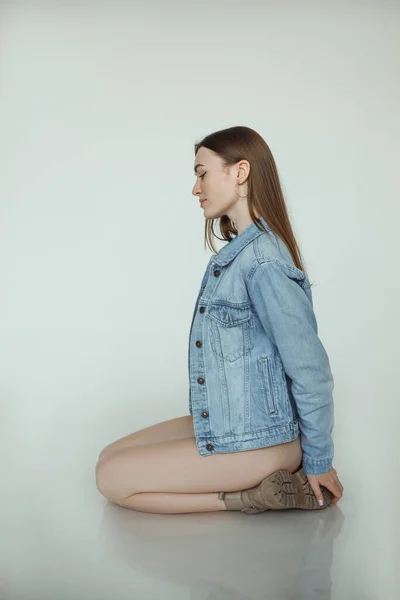 Vertical of seductive woman in jeans jacket, sit on bend naked legs on the floor, white background. Enjoying her body. Stylish lady posing. Healthy lifestyle, sport, body care. Side view. Copy space