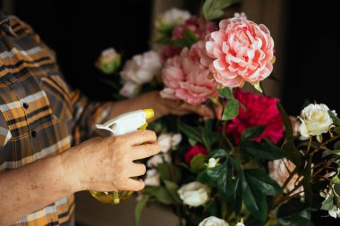 Üst düzey bir kadının kareli gömlek giydiği, pembe buket püskürttüğü, sarı tozluklu beyaz güller sıktığı, çiçeklere dokunduğu kesilmiş bir fotoğraf. Çiçekçi, botanik, anneler günü, kadınlar günü, evlilik yıldönümü.. 