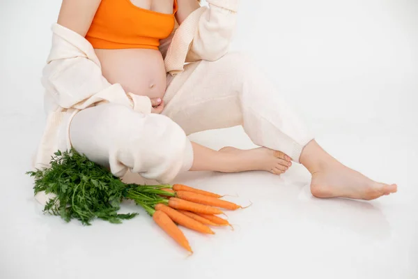 被裁剪的孕妇的照片 她拿着一捆橘子蔬菜 绿色的胡萝卜坐在一起 穿着亚麻衣服 小心地握住腹部 新鲜食品营养饮食 健康饮食 白人背景 — 图库照片