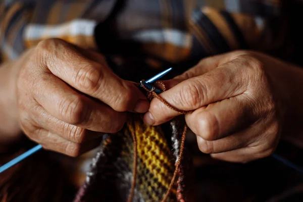 Close-up of hands of unrecognizable senior woman wearing checkered shirt, knitting colorful scarf with blue metal needles, brown yarn. Needlework, leisure activities, handiwork, handicraft, hobby.