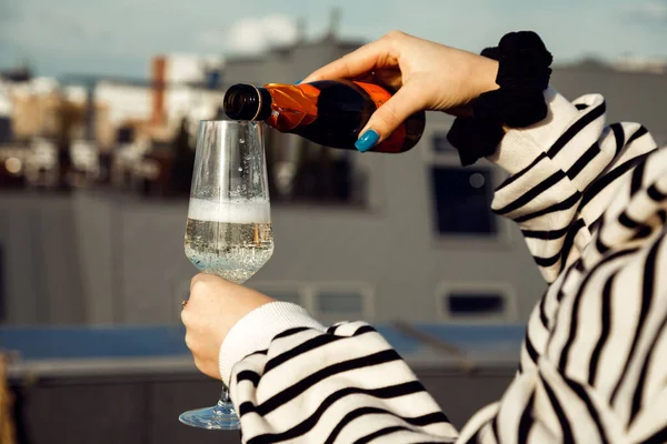 Cropped photo of woman wearing black and white striped sweatshirt, black scrunchy for hair on hand, pouring champagne from small brown bottle into glass on city background.