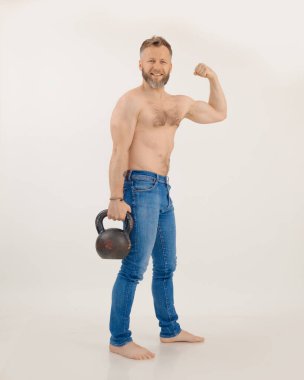 Çıplak gövdeli genç atletik adam elinde kettlebell ve gergin kaslarıyla beyaz arka planda duruyor. Güç egzersizi, ağırlık kaldırma, kasları şişirme. Spor ve sağlıklı yaşam tarzı kavramı.