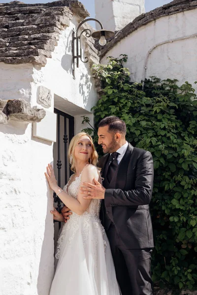 白いドレスを着たブロンドの花嫁とスーツを着たブルネットの新郎の肖像画は イタリアの街の通りでお互いを受け入れ 古い建物の背景 美しくロマンチックな結婚式幸せなカップル — ストック写真
