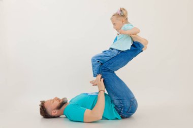 Kolları şişmiş genç atletik bir adam sırt üstü uzanıyor ve bacaklarını kaldırıyor küçük kız, beyaz arka plan. Bütün aile idmanı, baba kız. Spor ve sağlıklı yaşam tarzı kavramı.