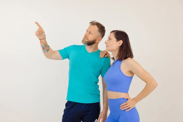 Young athletic woman and man in blue sportswear standing and pointing by finger, white background. Keeping fit by fitness workout in gym, pumping up body. Family sport and healthy lifestyle concept.
