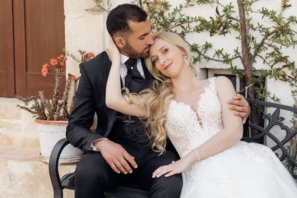 白いドレスを着た若いブロンドの花嫁とスーツを着たブルネットの新郎の肖像画は イタリアの旧市街の建物の近くのベンチに抱きしめ 座っています 美しくロマンチックな結婚式幸せなカップル — ストック写真