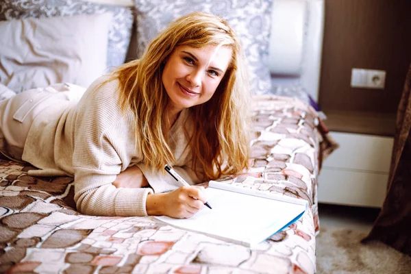 Smiling blonde woman in home clothes lying on bed and writing with pen in notebook closeup. Young happy lady relaxing at home in cozy bedroom in morning. Working and useful activity concept.