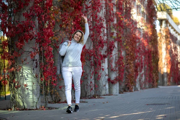 Elegant blonde in white trousers and pale blue cashmere sweater greets someone with wave of her hand walking through autumn city along wall covered with red girlish grapes. Walking in fresh air.