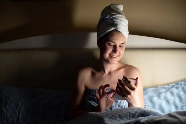 白いタオルを頭の周りにベッドの毛布で覆われて横に包まれた若い笑顔の女性の肖像画は スマートフォンを見て 画面に触れると 入力します テクノロジー ソーシャルメディア ネットサーフィン — ストック写真