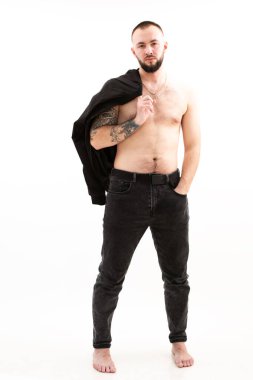 Kendine güvenen, havalı, seksi, sakallı ve dövmeli çekici bir adamın dikey portresi. Beyaz arka planda stüdyo resmiyle üstsüz ve üstsüz poz veriyor. Sıradan moda. Tüm vücut uzunluğu