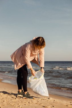 Kadın gönüllü ve asistanı, plastikten ve çöpten deniz kenarındaki kumsalın temizlenmesine yardım ediyor. Poşetlemek için çöp topluyorum. Rüzgarlı bir hava. Ekolojik geri dönüşüm sorunu, ekosistemi korumak