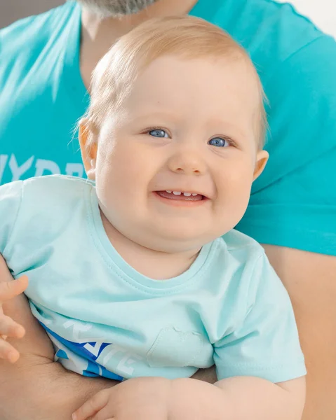 Portrait of family wearing blue T-shirts. Little smiling blue-eyed baby toddler child with short fair hair sitting on man. Father holding son. Family, love, care, relationship, parenthood. Vertical.